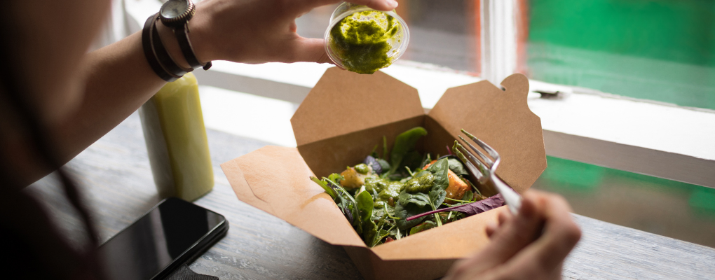 Comida para llevar en caja de cartón compostable con el logotipo de Restaurante sostenible, siendo aderezada con pesto verde fresco.